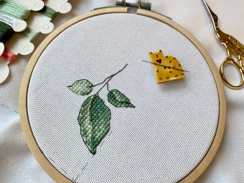 cross stitch piece with leaves stitched on and a cat needle holder resting on the piece